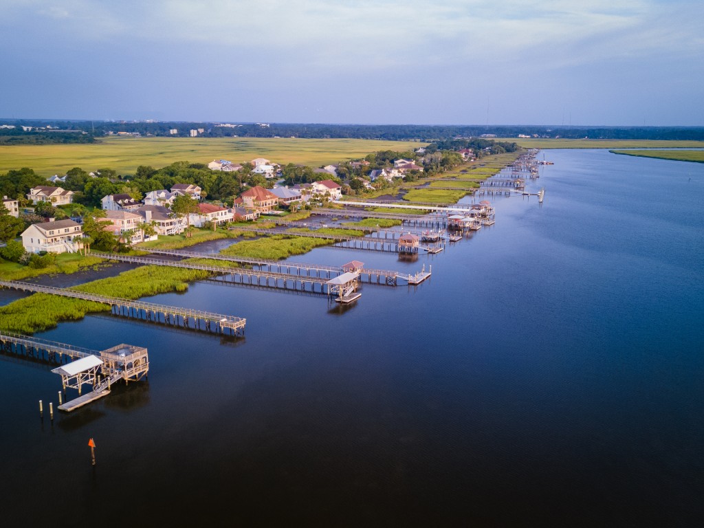 Aerial view of a tranquil waterway lined with docks and residential homes, surrounded by lush green marshland under a soft blue sky.