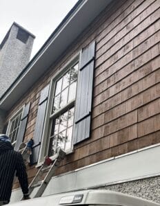Painter working on dark board-and-batten shutters on a wood shingle home