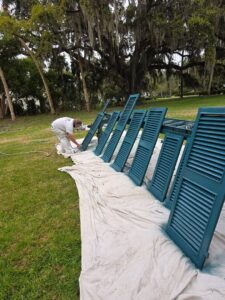 Painter spraying teal louvered shutters outdoors during exterior refinishing