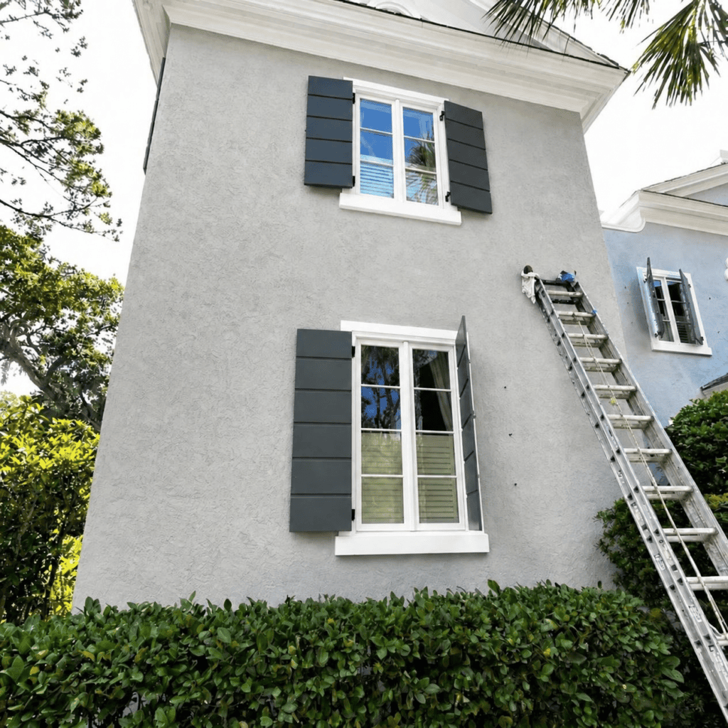 Sea Palms House Painters Gray stucco exterior with dark shutters and a ladder against the wall during painting