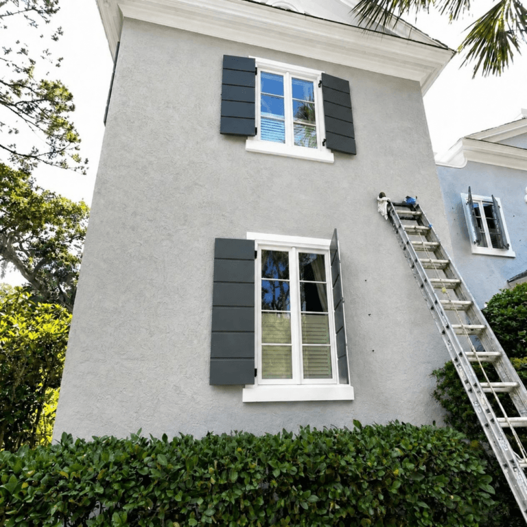 Gray stucco exterior with dark shutters and a ladder against the wall during painting